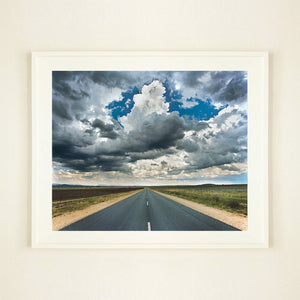 White framed photograph by Richard Heeps. A photograph looking along a long straight road into the distance, it is edged either side with sand and then prairie land. The vast, cloudy sky sits above.