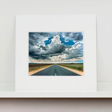 Load image into Gallery viewer, Mounted photograph by Richard Heeps. A photograph looking along a long straight road into the distance, it is edged either side with sand and then prairie land. The vast, cloudy sky sits above.