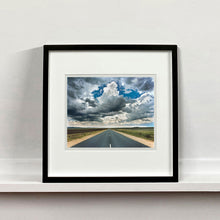 Load image into Gallery viewer, Black framed photograph by Richard Heeps. A photograph looking along a long straight road into the distance, it is edged either side with sand and then prairie land. The vast, cloudy sky sits above.