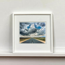 Load image into Gallery viewer, White framed photograph by Richard Heeps. A photograph looking along a long straight road into the distance, it is edged either side with sand and then prairie land. The vast, cloudy sky sits above.