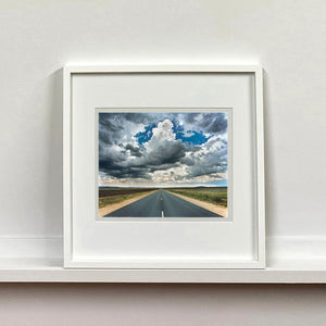 White framed photograph by Richard Heeps. A photograph looking along a long straight road into the distance, it is edged either side with sand and then prairie land. The vast, cloudy sky sits above.