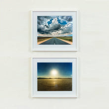 Load image into Gallery viewer, Two white framed photographs by Richard Heeps. The top one is a photograph looking along a long straight road into the distance, it is edged either side with sand and then prairie land. The vast, cloudy sky sits above. The photo below is a vast sky with a hazy sun shining and below is a flat beige ground with circular marks on it.