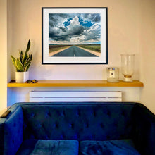 Load image into Gallery viewer, In situ photograph by Richard Heeps. A photograph looking along a long straight road into the distance, it is edged either side with sand and then prairie land. The vast, cloudy sky sits above.