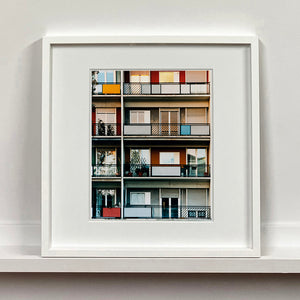 White framed photograph by Richard Heeps. This is the balconies and balcony doors of 4 floors of flats located on the Rue Dezza. The colours of the walls alternate between red and grey but look different as the light catches them.