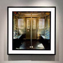 Load image into Gallery viewer, Black framed photograph by Richard Heeps. Photograph of the inside of a subway car, looking towards the coaches through the emergency adjoining door. A grab pole sits in the middle.