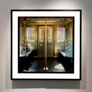 Black framed photograph by Richard Heeps. Photograph of the inside of a subway car, looking towards the coaches through the emergency adjoining door. A grab pole sits in the middle.