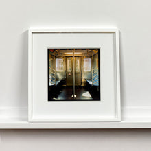Load image into Gallery viewer, White framed photograph by Richard Heeps. Photograph of the inside of a subway car, looking towards the coaches through the emergency adjoining door. A grab pole sits in the middle.
