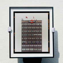 Load image into Gallery viewer, Black framed photograph held by photographer Richard Heeps. High rise offices with Martini logo on the top facade. 