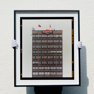 Black framed photograph held by photographer Richard Heeps. High rise offices with Martini logo on the top facade. 