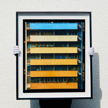 Load image into Gallery viewer, |A black framed photograph held by photographer Richard Heeps. Photograph of an apartment building with coloured balconies, blue at the top balcony and then fading from light yellow to a sunburnt yellow at the bottom.