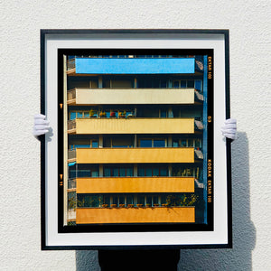 |A black framed photograph held by photographer Richard Heeps. Photograph of an apartment building with coloured balconies, blue at the top balcony and then fading from light yellow to a sunburnt yellow at the bottom.