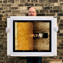 Load image into Gallery viewer, Photograph held by photographer Richard Heeps. Brown flecked marble walls in different tones. In the middle is half a brown plaque with golden letters showing half an A, followed by a D and an A.