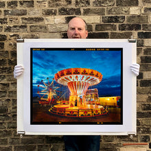 Load image into Gallery viewer, Photograph held by the photographer Richard Heeps. A fairground ride, the chairoplanes, sits lit in golden and red colours against a dark blue sky.