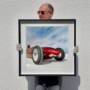 Black framed photograph held by photographer Richard Heeps.  The side on front of a red belly tank racing car, it is designed as a shark eating a $100 bill.