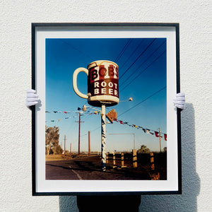 Photograph held by photographer Richard Heeps. A giant model of a mug with Bob's Root Beer written on it sits on top of a giant pole. There is bunting hanging from the pole. It sits alongside a power line on a remote looking American country road.