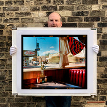 Load image into Gallery viewer, Photograph held by photographer Richard Heeps. Inside an American diner the light is shining on the table and the seat is the classic vibrant red faux-leather of a retro diner. The photograph is looking out of the window into the outside street.