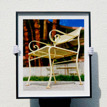 Load image into Gallery viewer, Photograph by Richard Heeps. A cream chair sits on hard standing, behind the chair and slightly out of focus is lush green grass and warm red tree trunks.