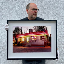 Load image into Gallery viewer, Black framed photograph held by Richard Heeps. This photograph depicts a one storey small building &quot;Dot&#39;s Diner&quot; brightly lit with a pink roof, with Hamburgers, Hot Dogs, Shakes, Fries written along the top width of the building.