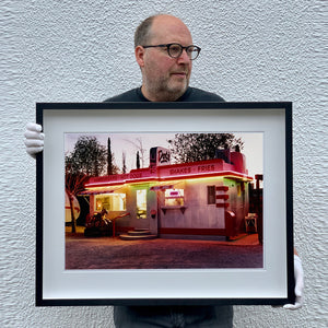 Black framed photograph held by Richard Heeps. This photograph depicts a one storey small building "Dot's Diner" brightly lit with a pink roof, with Hamburgers, Hot Dogs, Shakes, Fries written along the top width of the building.
