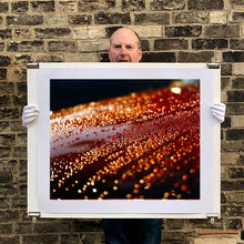 Load image into Gallery viewer, Photograph held by photographer Richard Heeps. The photograph is of a brown metal surface with water droplets on. The lighting makes the droplets appear in different colours of orange and brown.
