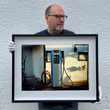 Load image into Gallery viewer, Black framed photograph held by Richard Heeps. A vintage petrol pump with a white front and blue sides, sitting outside a white slatted building.