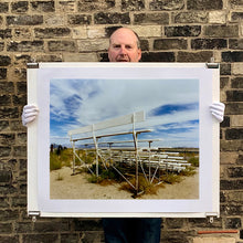 Load image into Gallery viewer, Photograph held by photographer Richard Heeps. The back view of a basic white painted grandstand sits in the middle of this photograph. Grass grows up from its base and it sits alone on a sandy ground.