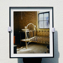 Load image into Gallery viewer, Black photograph held by photographer Richard Heeps. A run down now unused room with the metal surround of a cot bed and no mattress, at the end of the bed a wash stand with a bowl on top. Light shines in the room from the window.