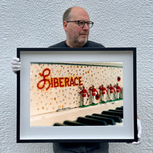 Load image into Gallery viewer, Black framed photograph held by the photographer, Richard Heeps. A close up of Liberace&#39;s piano. This captures the black keys and the fall board which is decorated with crystals and red buttons spelling out Liberace in capitals. There are also 5 red and white metal soldiers positioned marching along the rim.