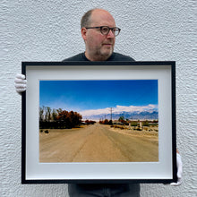 Load image into Gallery viewer, Black framed photograph held by Richard Heeps. A dusty road in the middle, heading towards the snow capped mountains in the distance, on the right are brown bushes and trees and on the left, single level concrete buildings.