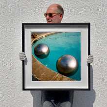 Load image into Gallery viewer, Black framed photograph held by photographer  Richard Heeps. The corner of a circular swimming pool with two metallic silver beach balls floating on the water.