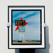 Load image into Gallery viewer, Photograph held by photographer Richard Heeps edged by film rebate. A roadside sign on Route 66 in America. The word ROY&#39;S appears in a black sign with a bit red arrow pointing to the left ground, below this VACANCY and on a green square the words MOTEL and CAFE.