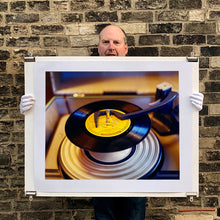 Load image into Gallery viewer, Photograph held by photographer Richard Heeps. A black vinyl with a yellow label sits high on the spindle of a deck under the overarm.