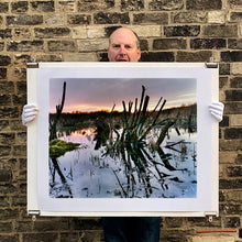 Load image into Gallery viewer, Photograph held by photographer Richard Heeps. Photograph of cut down, lichen clad branches poking out of the flooded fen field. The branches are strikingly dark and create dark reflections with a golden sunset in the background.