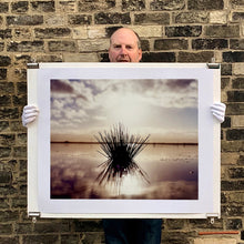 Load image into Gallery viewer, Photograph held by photographer Richard Heeps. A tussock is central to this photograph, black and reflected black into the fenland water below. The sky behind is dusky and atmospheric.