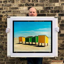 Load image into Gallery viewer, Photograph held by photographer Richard Heeps. Yellow and green beach lockers stand on their wooden legs on the clear sandy beach.