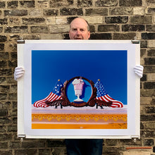 Load image into Gallery viewer, Photograph held by photographer Richard Heeps. A 3D shape milkshake parlour sign which has a pink milkshake with a white top, cherry and straws, surrounded by a wooden type shield, and on either side the look of draped American flags. This is set against a blue sky.
