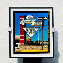 Load image into Gallery viewer, Black framed photograph by Richard Heeps. A group of signs on an American Road sitting outside a motel mounted on a post with a vertical line of red squares.  The top of  the sign shows an arrow pointing to the left to Pink Champagne, there is an arrow pointing down with MOTEL written in it. In front of this motel sign is a one way sign pointing to the right. 