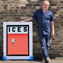 Load image into Gallery viewer, Photograph by Richard Heeps.  Richard Heeps holds a white framed print. At the top of the print, black letters spell out ICES and below is depicted a 99 icecream cone sitting left of centre against a melondrama red orange coloured background.  