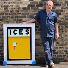 Load image into Gallery viewer, Photograph by Richard Heeps.  Richard Heeps holds a white framed print. At the top of the print, black letters spell out ICES and below is depicted a 99 icecream cone sitting left of centre against a mustard yellow coloured background.  