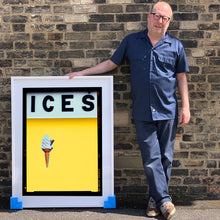 Load image into Gallery viewer, Photograph by Richard Heeps.  Richard Heeps holds a white framed print. At the top of the print, black letters spell out ICES and below is depicted a 99 icecream cone sitting left of centre against a sherbert yellow coloured background.  