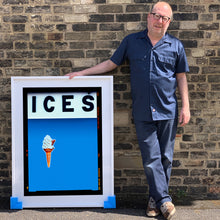 Load image into Gallery viewer, Photograph by Richard Heeps.  Richard Heeps holds a white framed print. At the top of the print, black letters spell out ICES and below is depicted a 99 icecream cone sitting left of centre against a sky blue coloured background.  