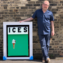 Load image into Gallery viewer, Photograph by Richard Heeps.  Richard Heeps holds a white framed print. At the top of the print, black letters spell out ICES and below is depicted a 99 icecream cone sitting left of centre against a viridian green coloured background.  