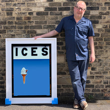 Load image into Gallery viewer, Photograph by Richard Heeps.  Richard Heeps holds a white framed print. At the top of the print, black letters spell out ICES and below is depicted a 99 icecream cone sitting left of centre against a baby blue coloured background.  