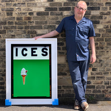 Load image into Gallery viewer, Photograph by Richard Heeps.  Richard Heeps holds a white framed print. At the top of the print, black letters spell out ICES and below is depicted a 99 icecream cone sitting left of centre against a green coloured background.  