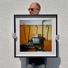 Load image into Gallery viewer, Black framed photograph held by photographer Richard Heeps. Photograph of a vintage room, a television is on a table with two aerials on top, beside the television are drinks and glasses. Hanging from the picture rail on the yellow wall behind the television are 4 different walking sticks and a bed pan. 