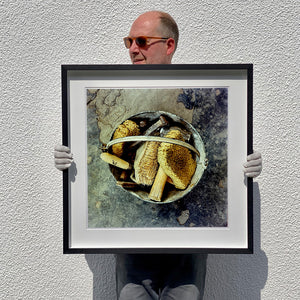 Black framed photograph held by photographer Richard Heeps. A bucket with sponges, brushes and wooden handled tools sit in a bucket on a cracked cement floor.