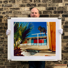 Load image into Gallery viewer, Photograph held by photographer Richard Heeps. A colourful but derelict vintage motel sits in the blue Californian sun. Palm trees appear behind and to the side. 