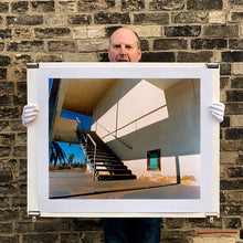 Load image into Gallery viewer, Photograph held by photographer Richard Heeps. The photograph has a metal staircase on the outside of a cream colour motel. The staircase has a ceiling but no sides so leads to blue sky. Behind the building is a blue sky and palm trees of the Californian Desert.