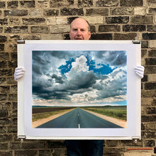 Load image into Gallery viewer, Photograph held by photographer Richard Heeps. A photograph looking along a long straight road into the distance, it is edged either side with sand and then prairie land. The vast, cloudy sky sits above.