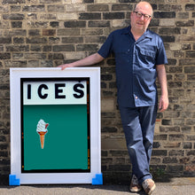 Load image into Gallery viewer, White framed photograph held by photographer Richard Heeps.  At the top black letters spell out ICES and below is depicted a 99 icecream cone sitting left of centre against a deep green teal coloured background.  